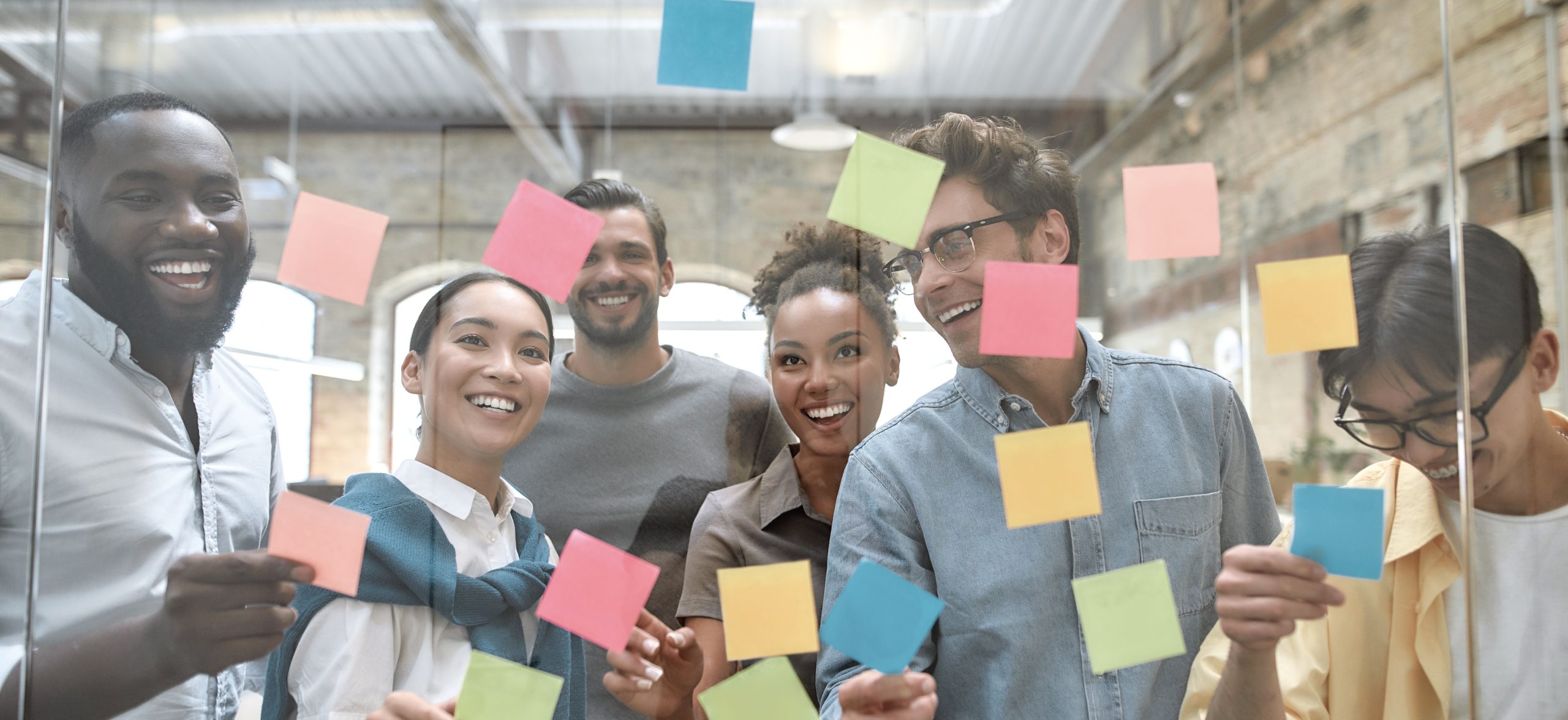 Working on business project together. Group of young and positive coworkers putting colorful sticky notes on a glass window in the creative office